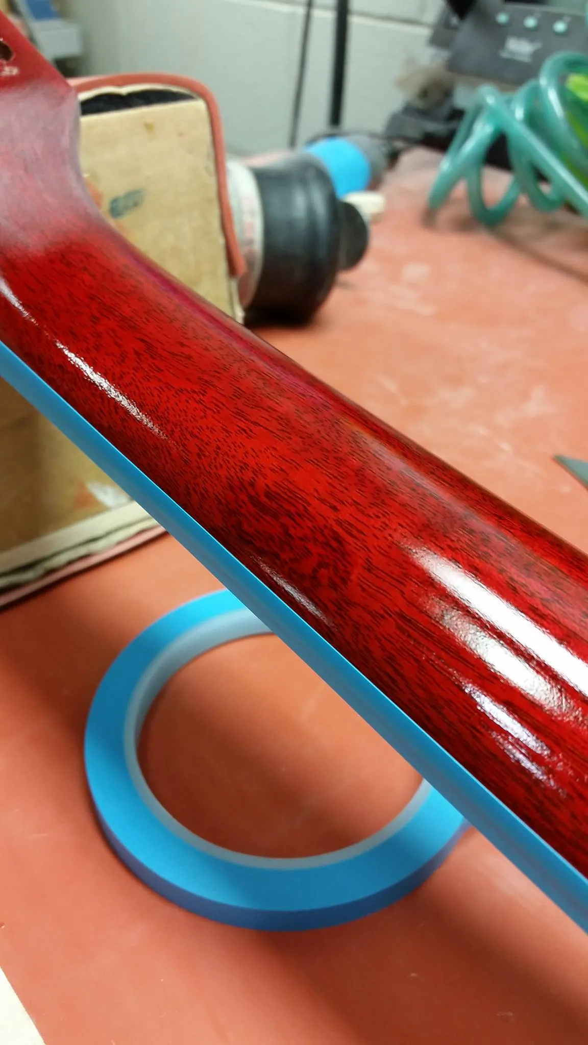 A cherry-red guitar neck with blue masking tape laid along the binding for finish work