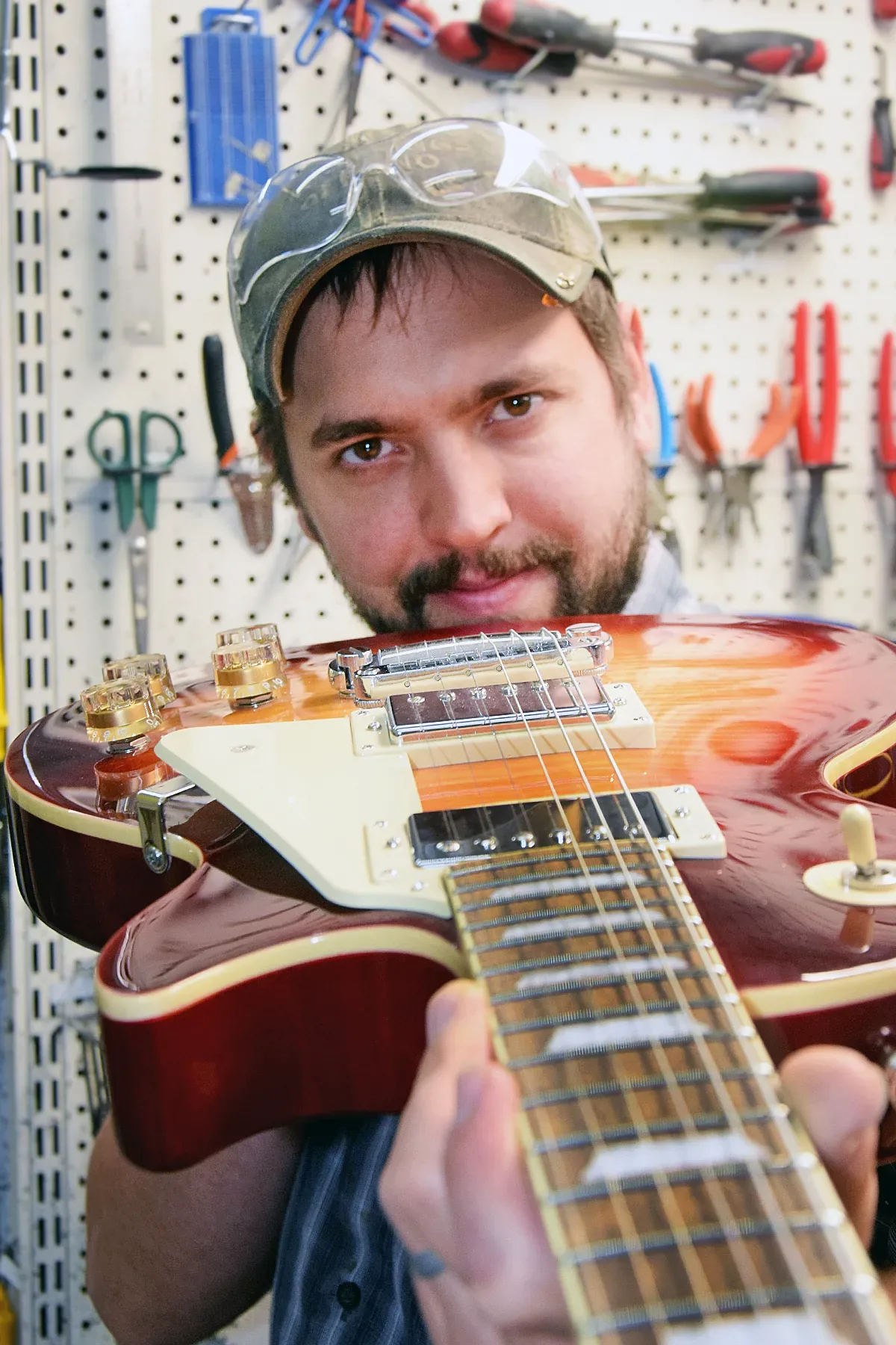 A luthier holding a cherry sunburst Les Paul–style guitar up to the camera in front of a pegboard of tools